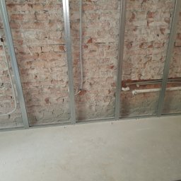 KoWbud - Surowa ceglana ściana z zamontowanymi metalowymi profilami, widoczne rury instalacji wodnej i elektrycznej, przygotowanie do dalszych prac wykończeniowych.