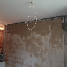 KoWbud - Ściana w trakcie szpachlowania, widoczne kable elektryczne pod sufitem i wiadro z zaprawą, ceglana ściana w tle