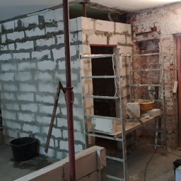 KoWbud - Wnętrze w trakcie prac murarskich: wznoszona ściana z bloczków, odsłonięta cegła na ścianach bocznych, drabina oparta o nową konstrukcję. Pracownik w kombinezonie i rękawicach murarskich...