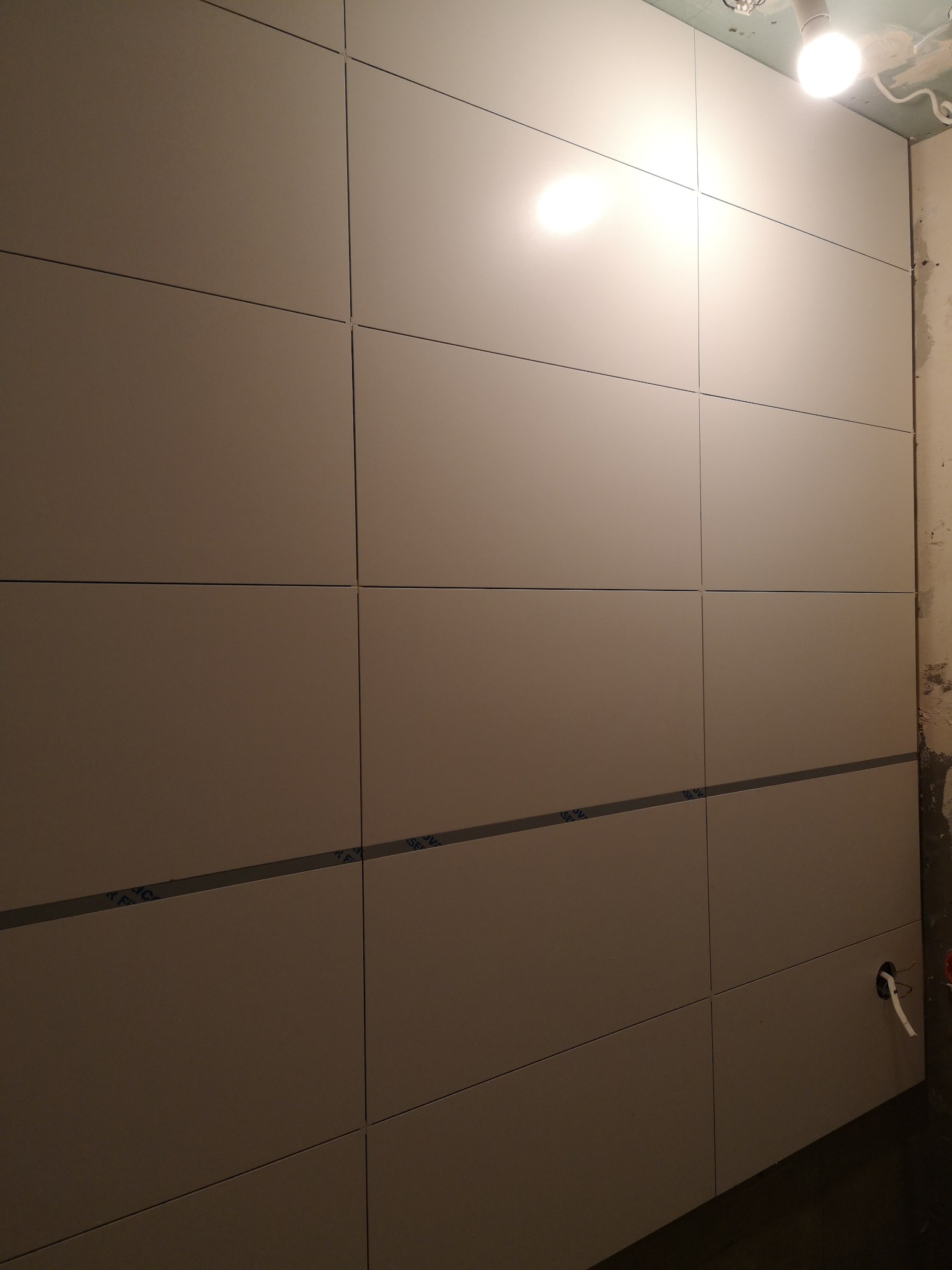Montaż wielkoformatowych płytek na ścianie w łazience, widoczne fugi i fragment instalacji elektrycznej przed wykończeniem.