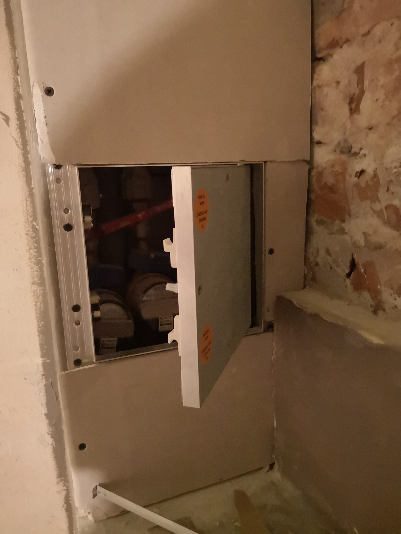 Otwarta rewizja wodomierzowa w trakcie montażu, widoczne rury i liczniki, obok fragment ściany z cegły.