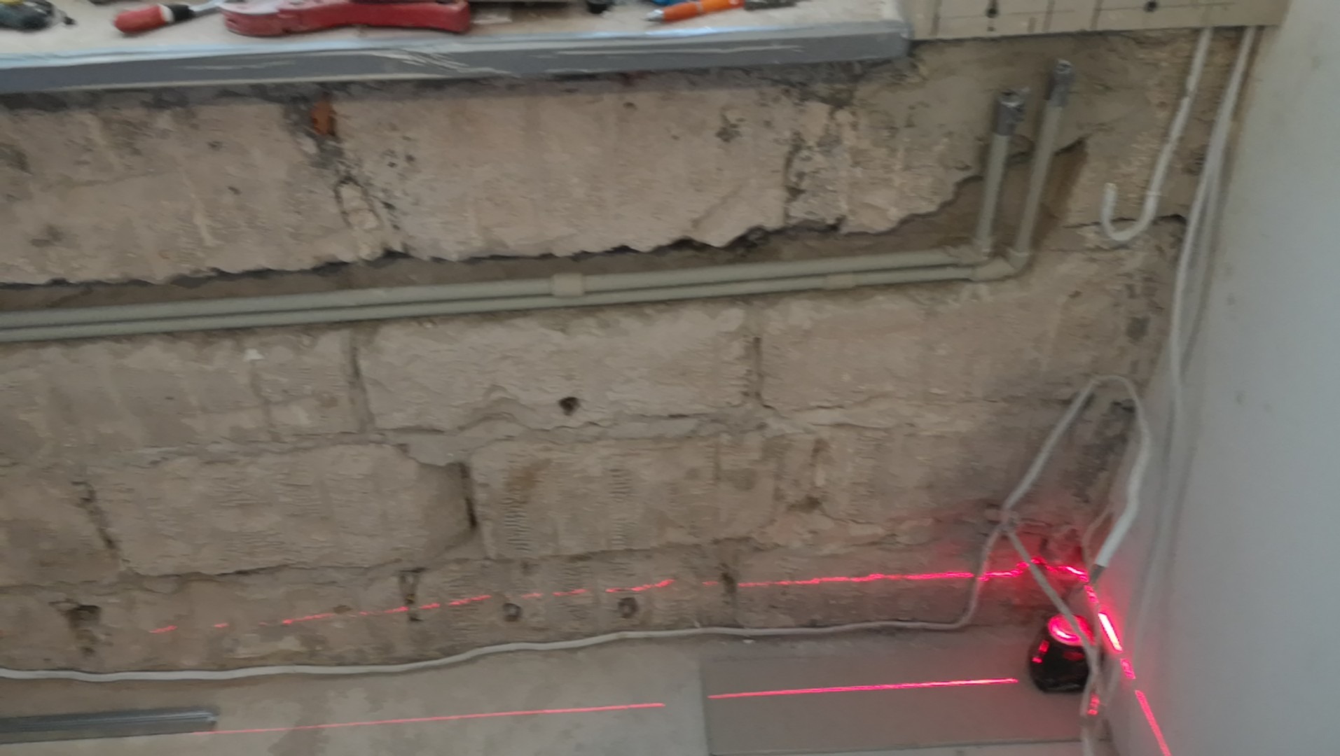 Surowa ściana z cegły i rurami, oświetlona czerwoną linią lasera, przygotowywana do prac wykończeniowych.