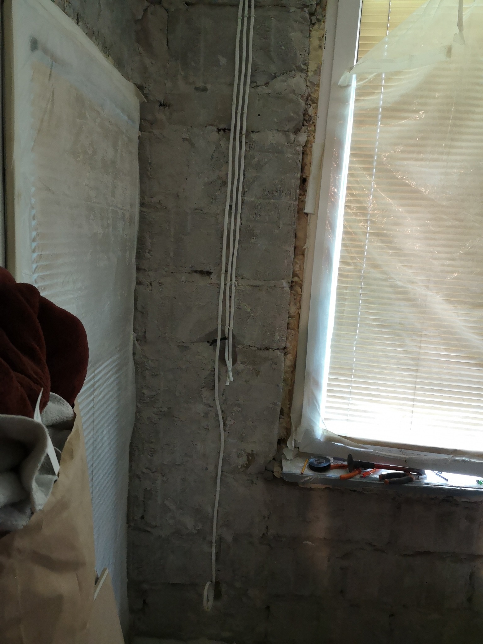 Ściana w trakcie przygotowania do prac wykończeniowych w Warszawie, widoczne przewody elektryczne, surowy tynk, okno zabezpieczone folią i narzędzia na parapecie.