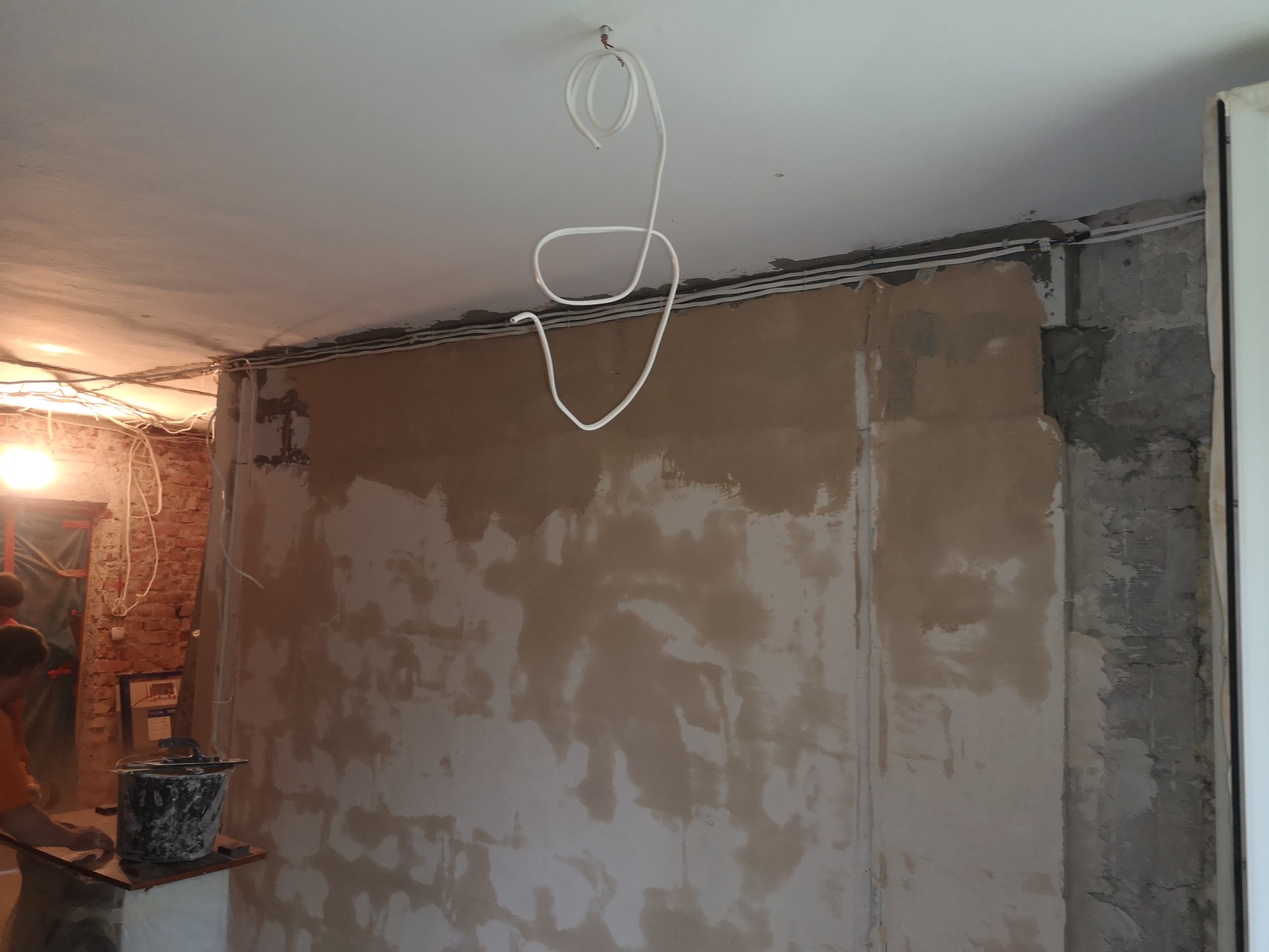 Ściana w trakcie szpachlowania, widoczne kable elektryczne pod sufitem i wiadro z zaprawą, ceglana ściana w tle