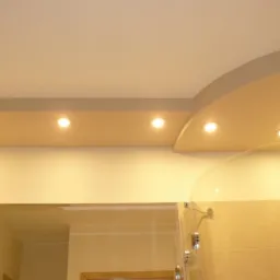 Podwieszany sufit w łazience z półokrągłym elementem i wbudowanymi punktowymi lampami halogenowymi, widoczny fragment kabiny prysznicowej z przezroczystymi drzwiami.