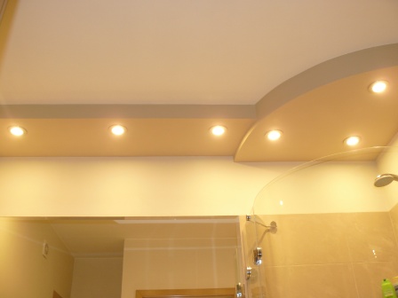 Podwieszany sufit w łazience z półokrągłym elementem i wbudowanymi punktowymi lampami halogenowymi, widoczny fragment kabiny prysznicowej z przezroczystymi drzwiami.