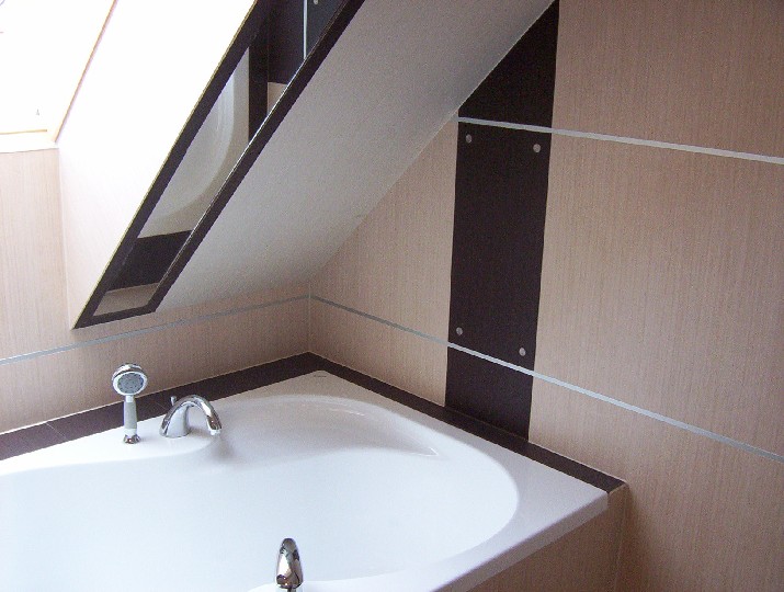 Biała narożna wanna z chromowaną armaturą i słuchawką prysznicową na tle beżowej ściany z ciemnobrązowymi pionowymi akcentami i srebrnymi listwami, widok na skos dachu.