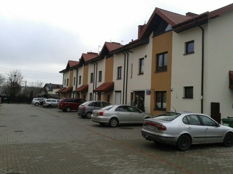 Szereg identycznych domów szeregowych z czerwonymi dachami i beżowo-brązowymi elewacjami, zaparkowane samochody na brukowanym parkingu.