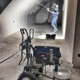 Malowanie natryskowe ścian w pomieszczeniu, pracownik w kombinezonie ochronnym i masce, widoczny agregat malarski z wyświetlaczem, surowy stan wykończenia wnętrza.
