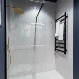 Nowoczesna łazienka z kabiną prysznicową typu walk-in, wykończona białymi, podłużnymi płytkami heksagonalnymi, złotą deszczownicą i czarnym grzejnikiem drabinkowym z białymi ręcznikami, na tle...