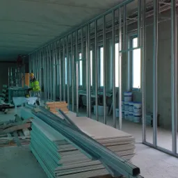 Montaż metalowej konstrukcji pod płytę karton gips w biurze, widoczny pracownik w żółtej kamizelce, stos płyt karton gips i profile, surowy beton na ścianach, okna w tle.