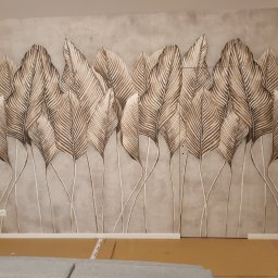 Repasbud - Ściana z naklejoną tapetą w motywie liści palmowych w odcieniach brązu i beżu na szarym tle, widoczne panele podłogowe i fragment sofy.