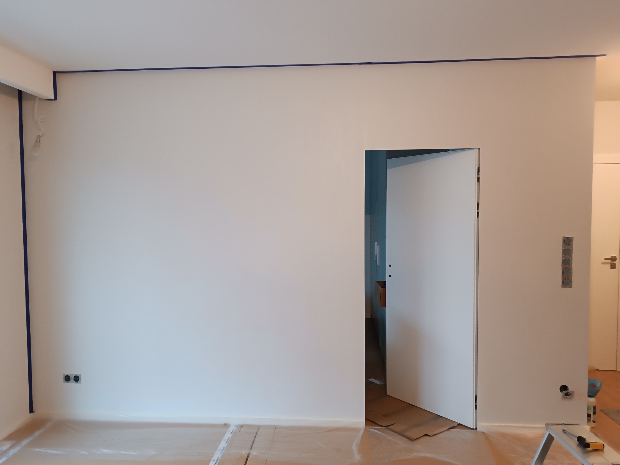 Przygotowanie do malowania ścian: zabezpieczona folią podłoga, obklejone taśmą malarską listwy i narożniki, widoczne gniazdka elektryczne i otwarta biała ściana.