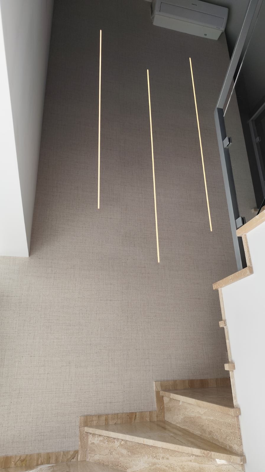 Beżowa ściana z fakturą tkaniny, oświetlona trzema pionowymi paskami światła LED, widoczne marmurowe schody i klimatyzator.