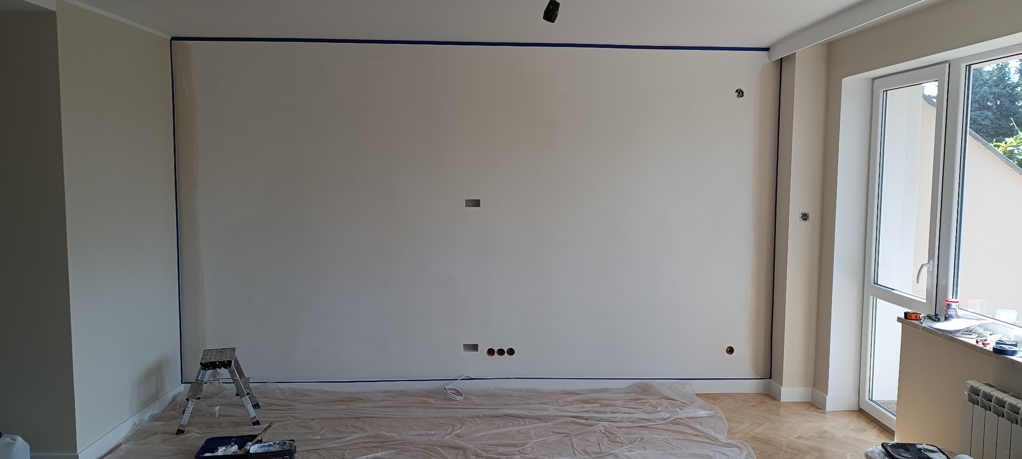 Przygotowana do malowania ściana z niezagruntowaną powierzchnią, zabezpieczona niebieską taśmą malarską na obrzeżach, z widocznymi gniazdkami elektrycznymi i osłoniętą folią podłogą, drabina...