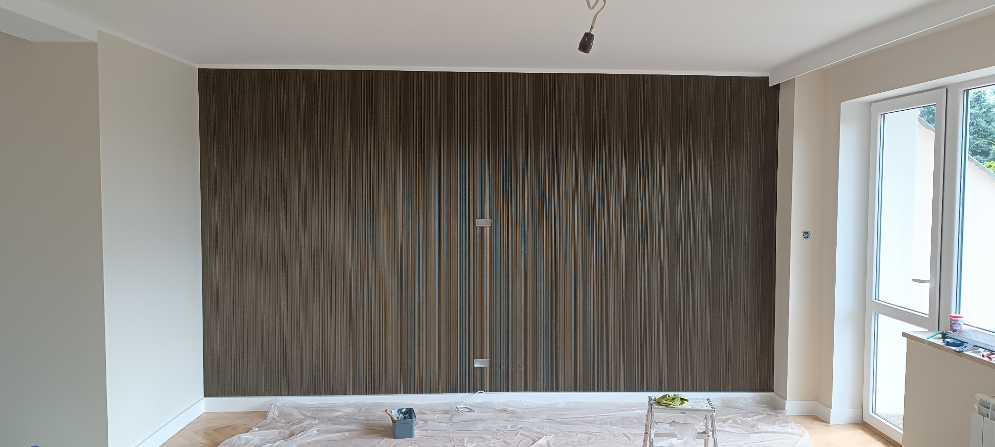 Ściana z brązową tapetą w pionowe prążki z elementami koloru niebieskiego i złotego, zabezpieczona folią malarską, widoczne gniazdka elektryczne i drabinka malarska.