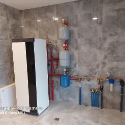 Nowoczesna kotłownia z białym piecem, zbiornikami wyrównawczymi i kompletną instalacją hydrauliczną na tle szarych płytek imitujących beton.