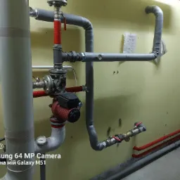 Węzeł instalacji hydraulicznej z pompą obiegową Grundfos, rurami izolowanymi szarą pianką i zaworami kulowymi z czerwonymi rączkami, na tle żółtej ściany z dokumentami.