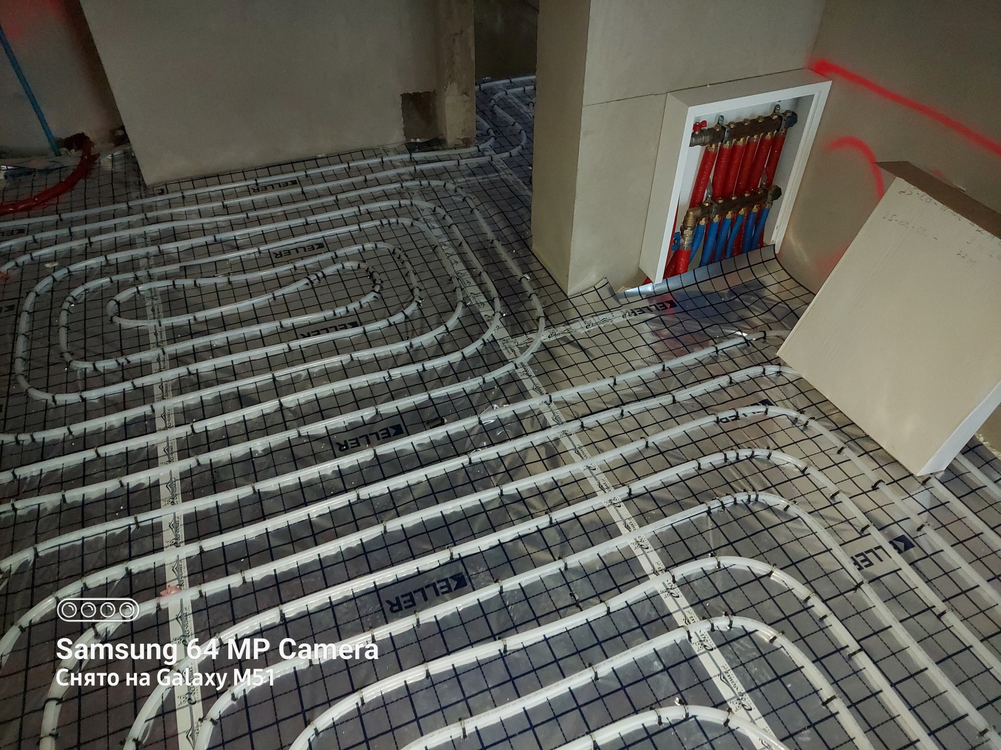 Rozłożona instalacja ogrzewania podłogowego z widocznym rozdzielaczem w szafce, rury w kolorach czerwonym i niebieskim, na podkładzie z folii z nadrukiem Keller.