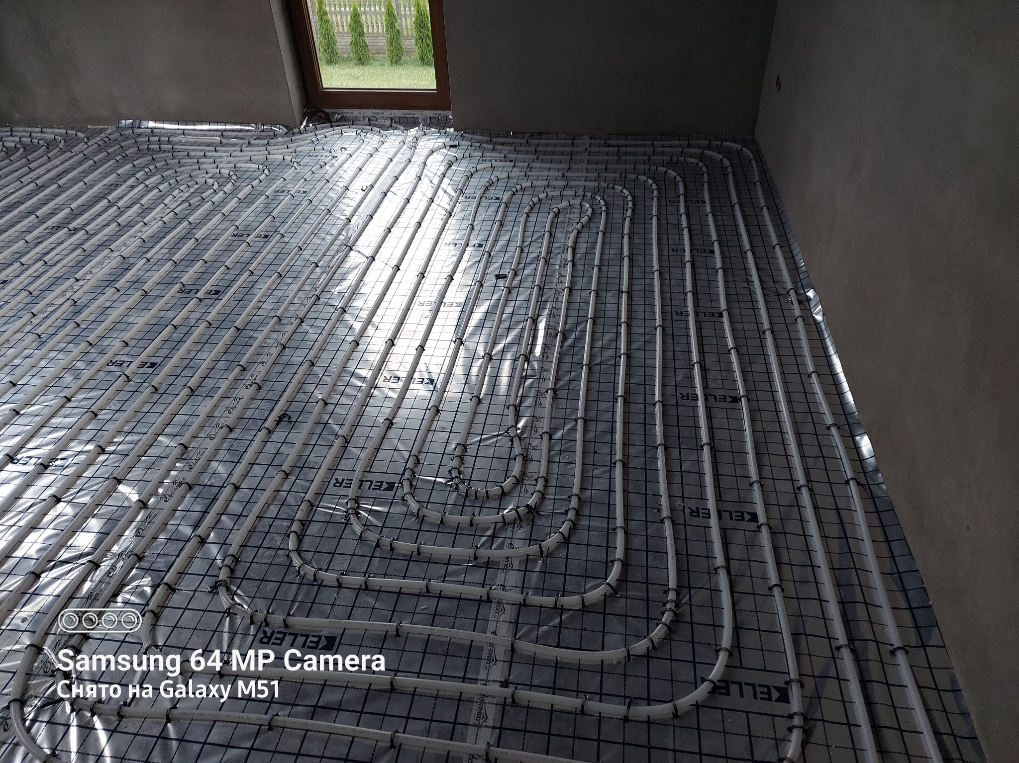 Białe rury ogrzewania podłogowego ułożone na folii z nadrukiem KELLER, widoczne na tle surowych ścian i okna z zielenią w tle.