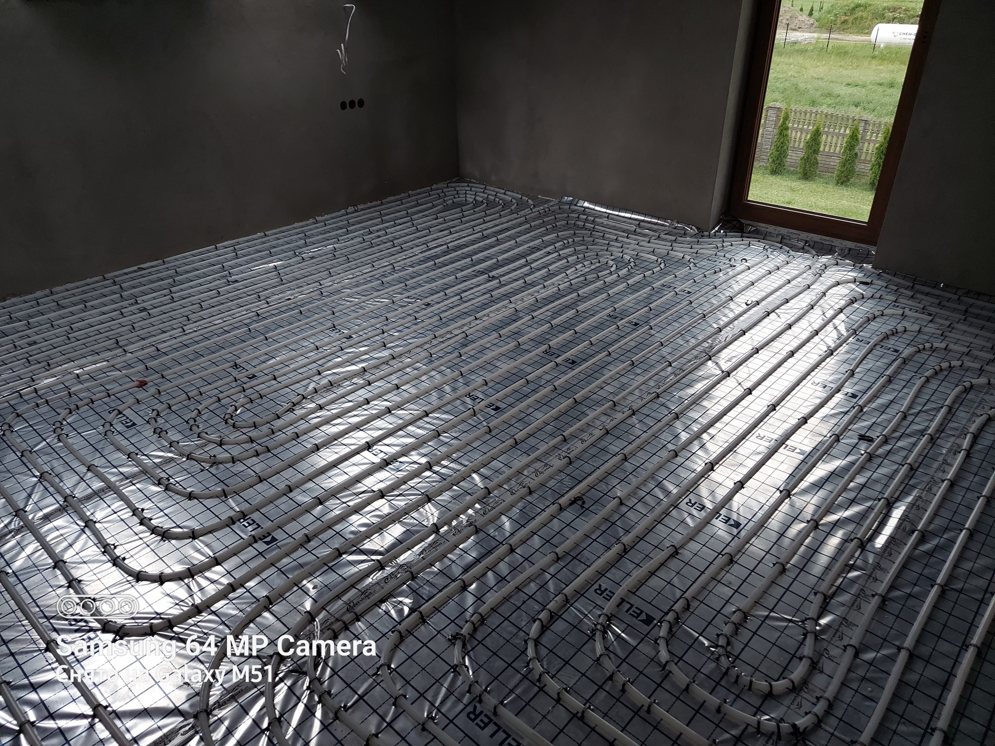 Rozłożone rury ogrzewania podłogowego na folii, widoczne elementy mocujące, surowe ściany w tle oraz okno z widokiem na zieleń.