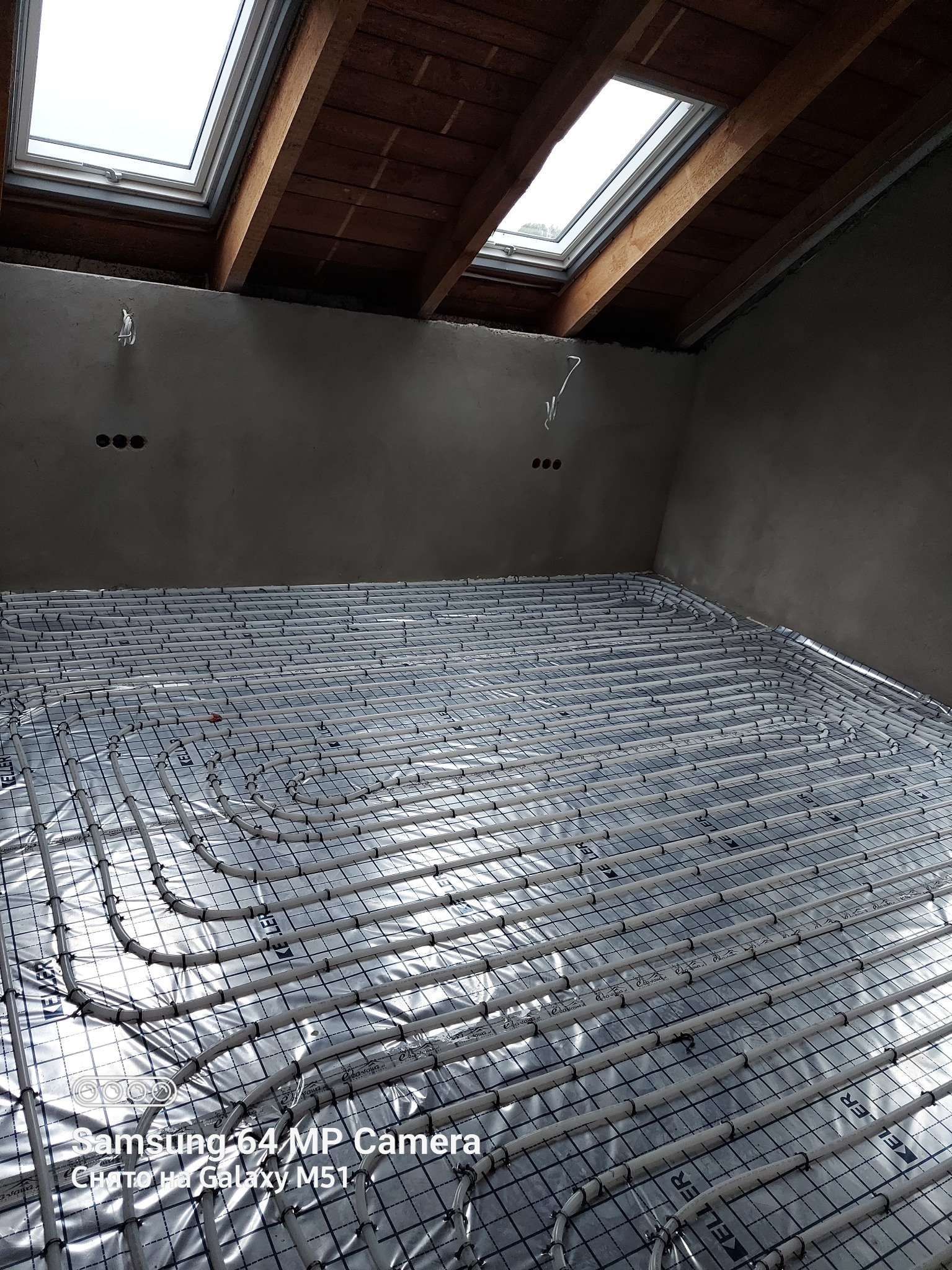 Rozłożone rury ogrzewania podłogowego na folii izolacyjnej z rastrem, widoczne okna dachowe i surowy stan ścian.