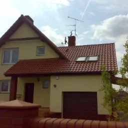 Żółty dom murowany z czerwoną dachówką, widoczny zza ceglanego murku, z anteną na kominie i drzewem po prawej stronie.