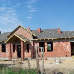 Dom murowany w trakcie budowy: widoczne ściany z czerwonej cegły, częściowo pokryty dach z papy i kominy, niezabudowane okna, paleta cegieł z przodu.