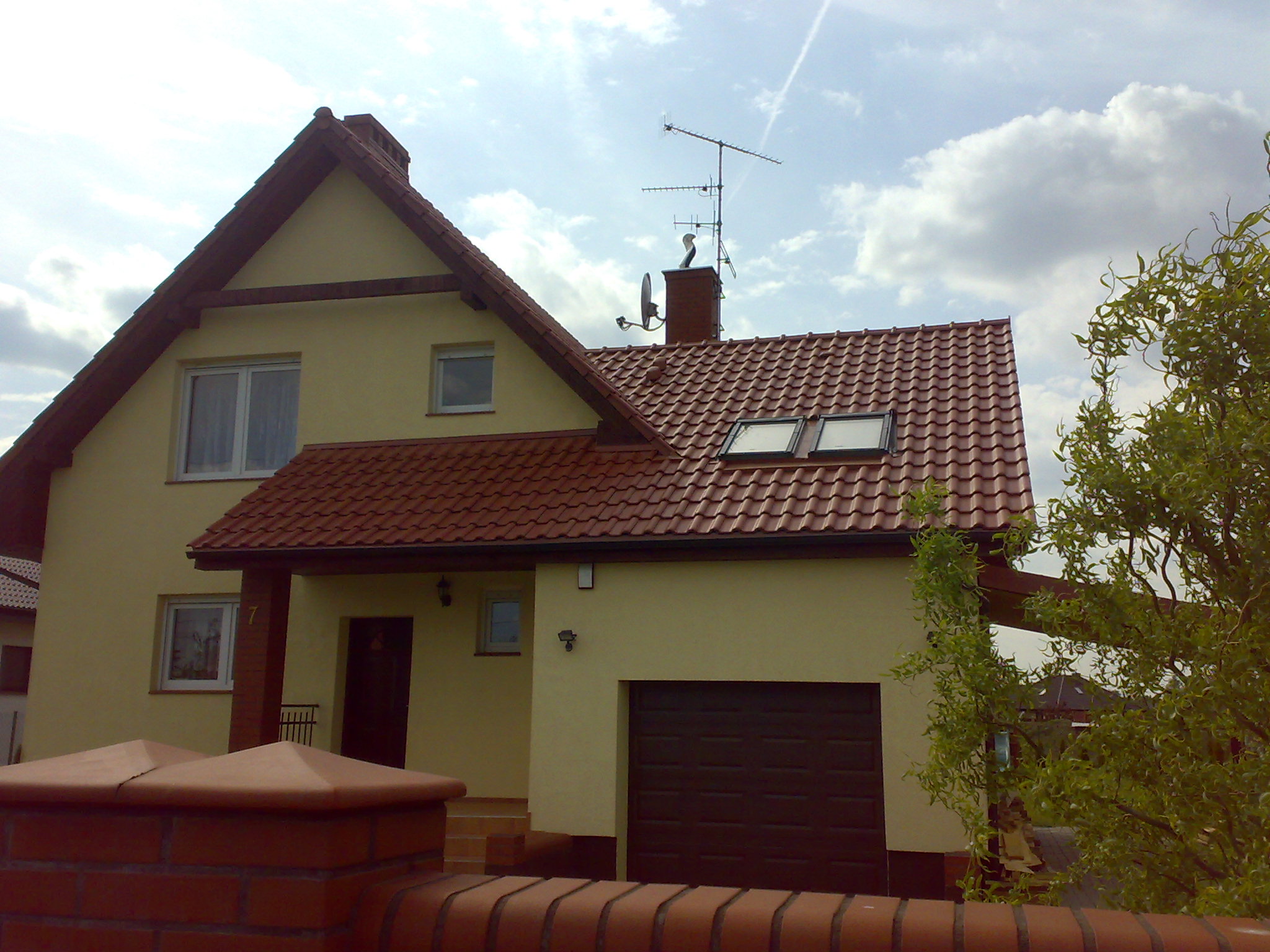 Żółty dom murowany z czerwoną dachówką, widoczny zza ceglanego murku, z anteną na kominie i drzewem po prawej stronie.