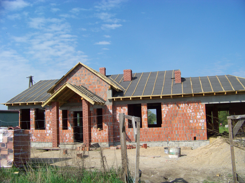 Dom murowany w trakcie budowy: widoczne ściany z czerwonej cegły, częściowo pokryty dach z papy i kominy, niezabudowane okna, paleta cegieł z przodu.