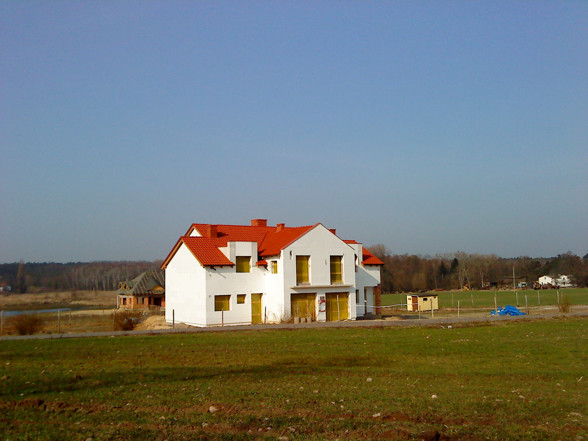 Nowy, dwupiętrowy dom murowany z czerwonym dachem w stanie surowym zamkniętym, z widocznymi żółtymi foliami na oknach i drzwiach, na tle zielonego pola i błękitnego nieba.