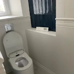 Nowoczesna toaleta myjąca z otwartą klapą, podświetlonym wnętrzem i panelem sterowania na tle ściany z ciemnoniebieskich, błyszczących płytek w wąskim formacie.