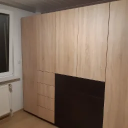 Duża szafa z frontami w kolorze jasnego drewna i ciemnymi szufladami, widoczna obok okna i grzejnika, na podłodze panele imitujące drewno.