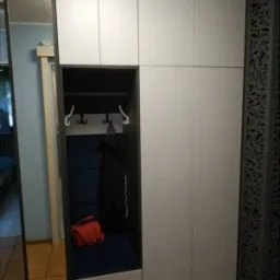 Zabudowa przedpokoju w Komornie z szafą z białymi frontami, siedziskiem z ciemnoniebieską tapicerką i wieszakami oraz dekoracyjnym panelem ażurowym.