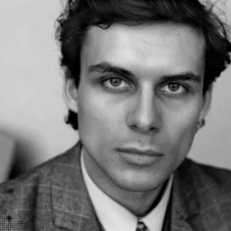 Czarno-białe portretowe ujęcie młodego mężczyzny w garniturze w kratę, z lekko rozczochranymi włosami i intensywnym spojrzeniem skierowanym wprost w obiektyw.