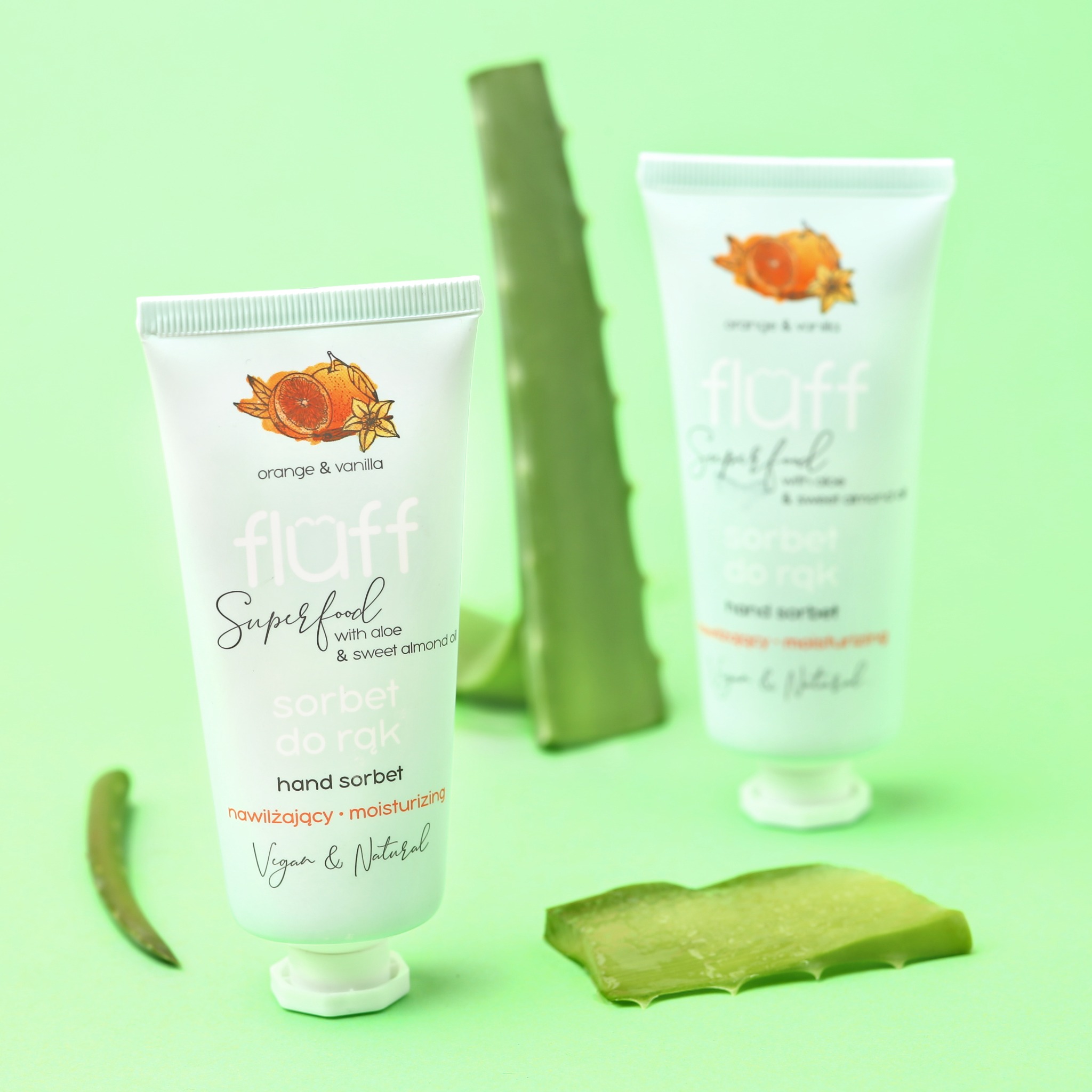 Dwie tubki kremu do rąk marki Fluff Superfood, o zapachu pomarańczy i wanilii, z dodatkiem aloesu, na tle kawałków świeżego aloesu i pastelowo zielonym tle.