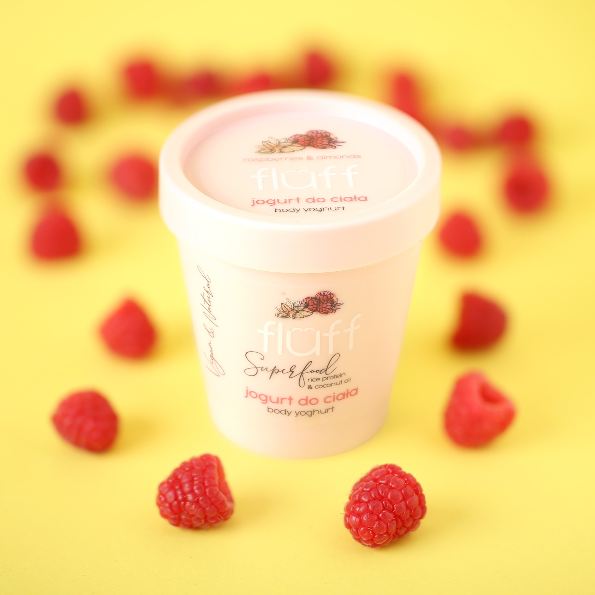 Packshot słoiczka z jogurtem do ciała marki Fluff na żółtym tle, otoczonego świeżymi malinami.