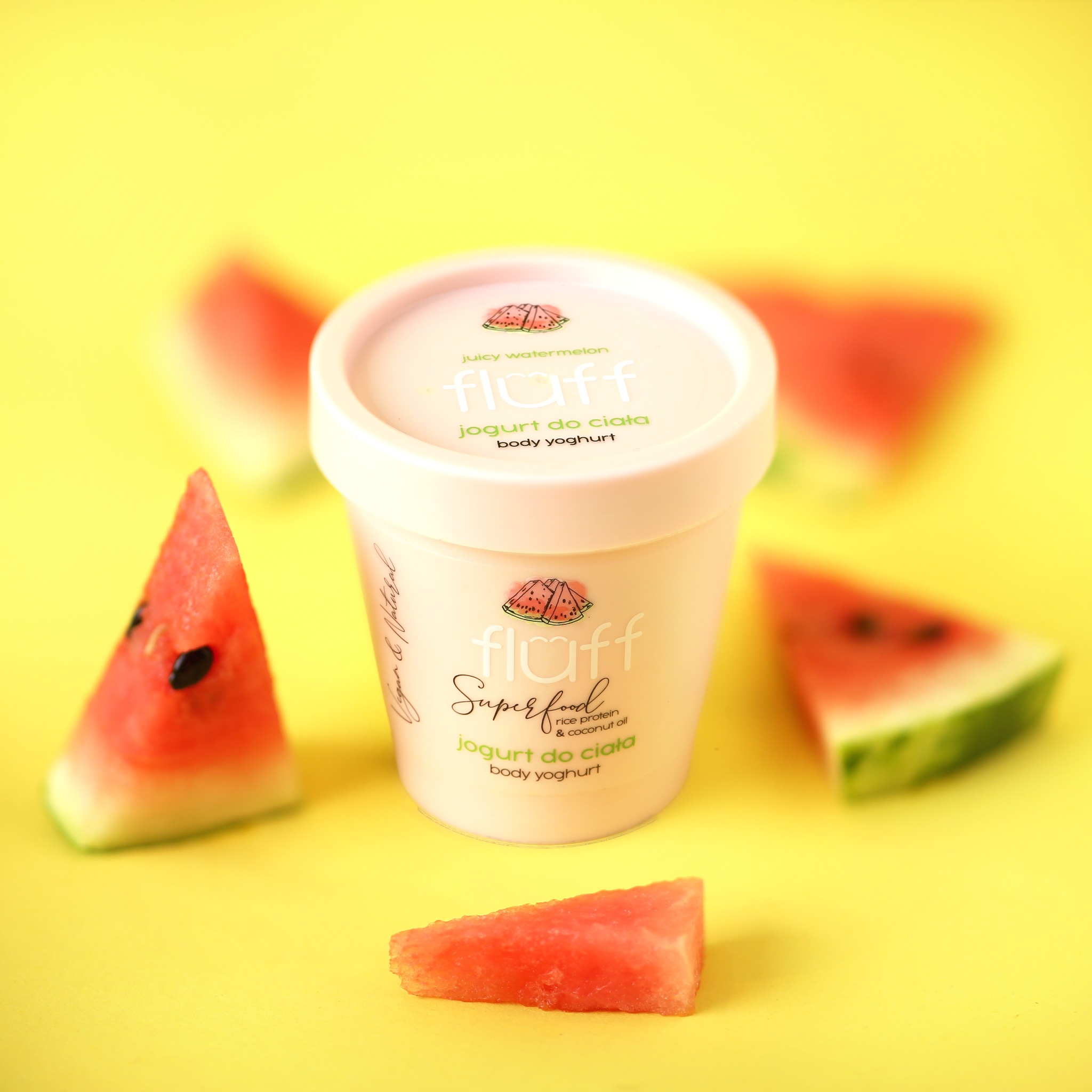 Packshot białego pojemnika jogurtu do ciała Fluff o zapachu arbuza, otoczonego kawałkami arbuza na żółtym tle.