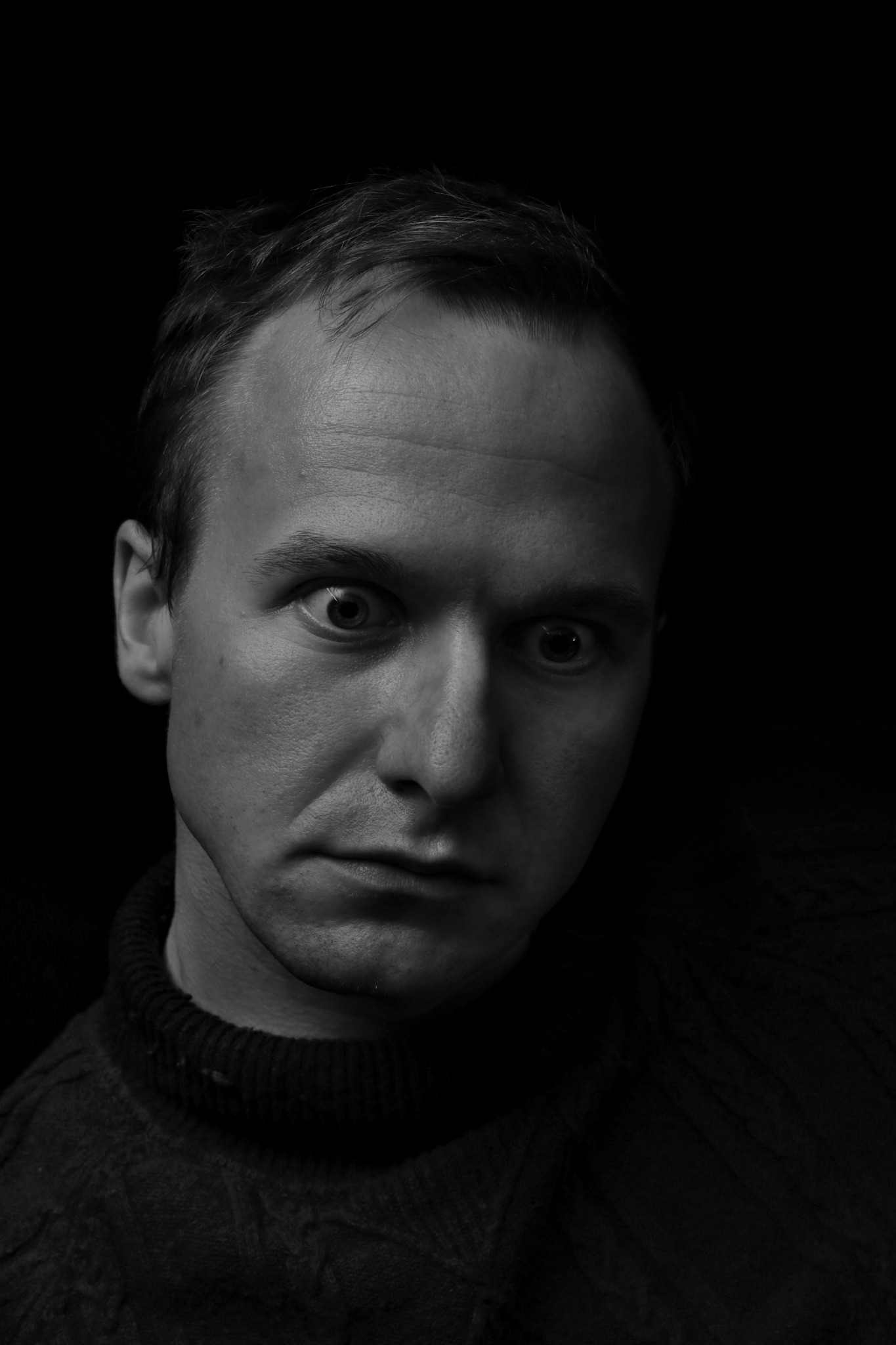 Czarno-biały portret mężczyzny z intensywnym spojrzeniem, na ciemnym tle, ubrany w sweter z golfem.