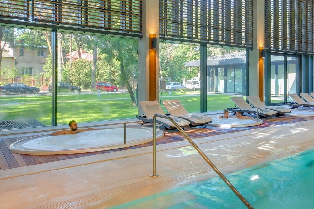 Wnętrze basenu solankowego z jacuzzi, leżakami i widokiem na zieleń za oknem, mężczyzna relaksuje się w wannie z hydromasażem.