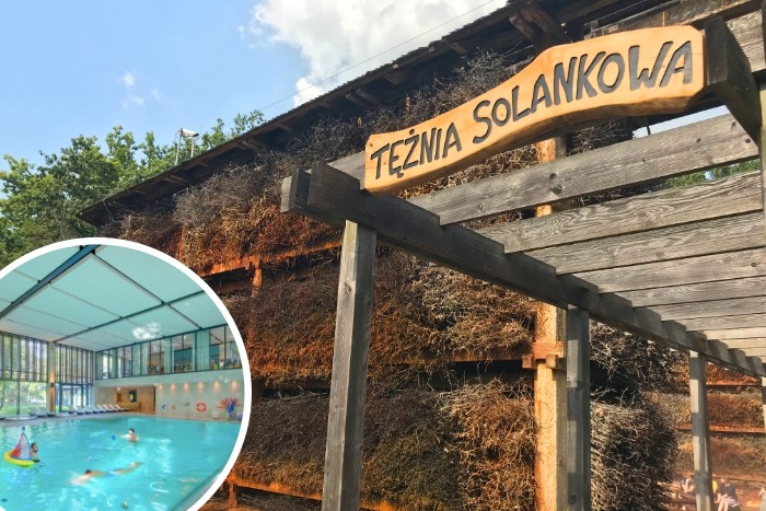 Tężnia solankowa z drewnianą konstrukcją i tablicą z nazwą, w tle basen kryty z pływającymi osobami, widoczne gałązki tarniny.