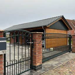Stalbau - Przesuwna brama wjazdowa z metalowymi prętami i dekoracyjnymi okręgami na tle drewnianego garażu z szarą dachówką, widok z zewnątrz posesji, skrzynka pocztowa z numerem domu po lewej.