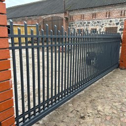 Stalbau - Grafitowa brama wjazdowa z pionowymi prętami i ozdobnymi zakończeniami, zamontowana pomiędzy ceglanymi słupkami na nierównej nawierzchni z kamienia i ziemi, na tle ceglanego budynku gospodarczego.