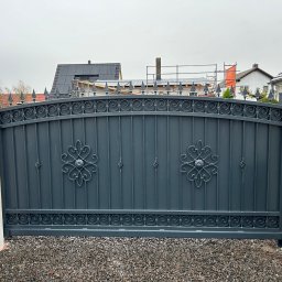 Stalbau - Szara brama wjazdowa z kutymi zdobieniami, osadzona między dwoma betonowymi słupkami na żwirowym podłożu, w tle zabudowa mieszkalna.