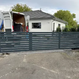 Nowoczesna, pozioma brama wjazdowa w kolorze szarym, częściowo zamontowana, z widocznym samochodem dostawczym firmy w tle oraz nowym domem jednorodzinnym.