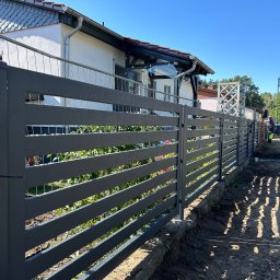 Stalbau - Nowoczesne, poziome ogrodzenie z szarych paneli metalowych, z widocznymi śladami po deszczu, na tle domu z białymi ścianami i zielenią ogrodu, z pracownikiem w tle.