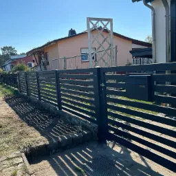 Długa, pozioma, ciemnoszara konstrukcja o regularnych prześwitach, oddzielająca posesję od chodnika, z widoczną skrzynką pocztową i otwartą furtką, w tle zabudowa jednorodzinna i błękitne niebo.