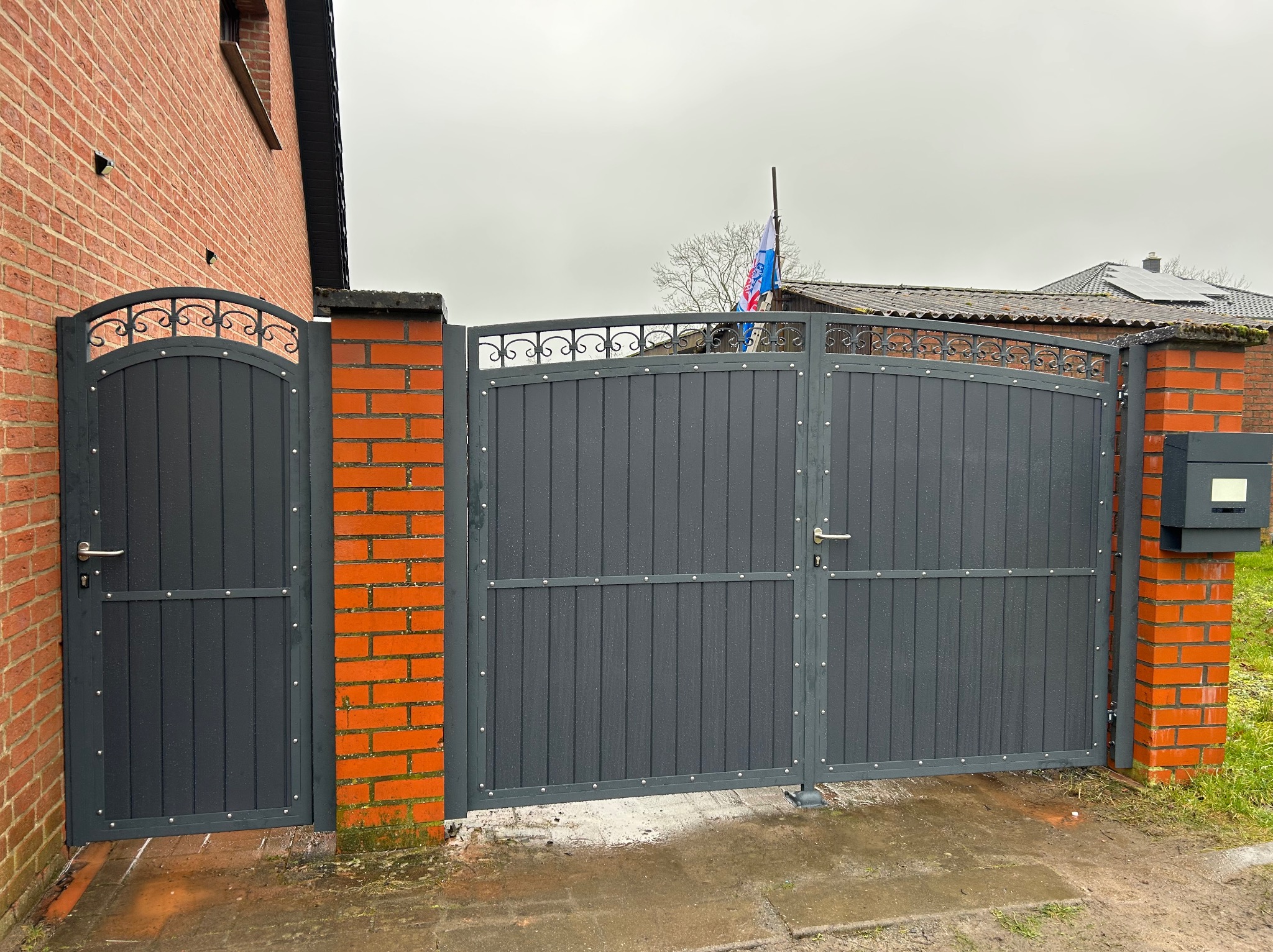 Szara brama wjazdowa i furtka z metalowymi panelami pionowymi, zdobionymi elementami kutymi u góry, osadzone na ceglanych słupkach, na mokrym podłożu.