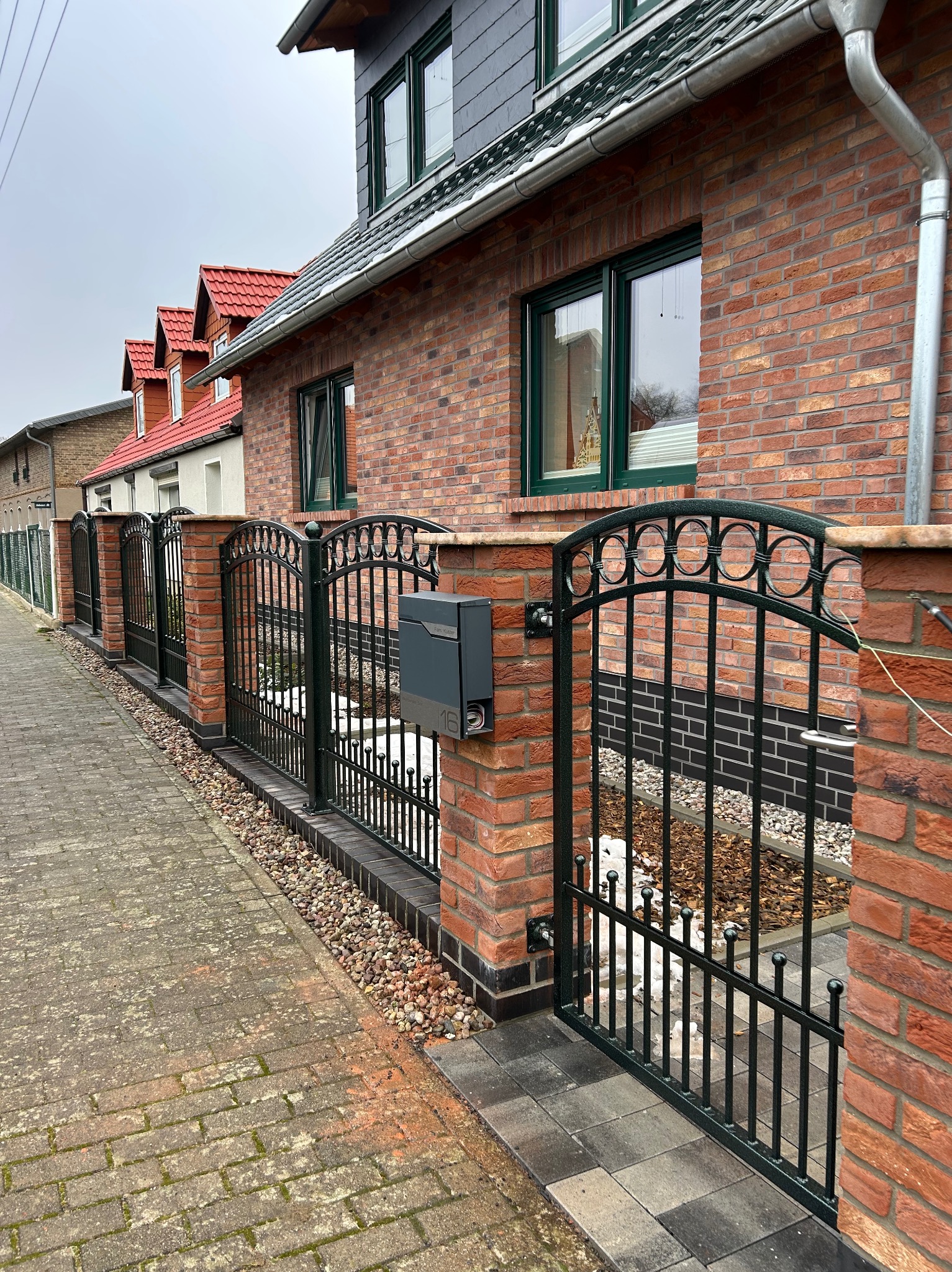 Czarne, kute przęsła ogrodzeniowe z ceglanymi słupkami i skrzynką na listy, widok z zewnątrz posesji.