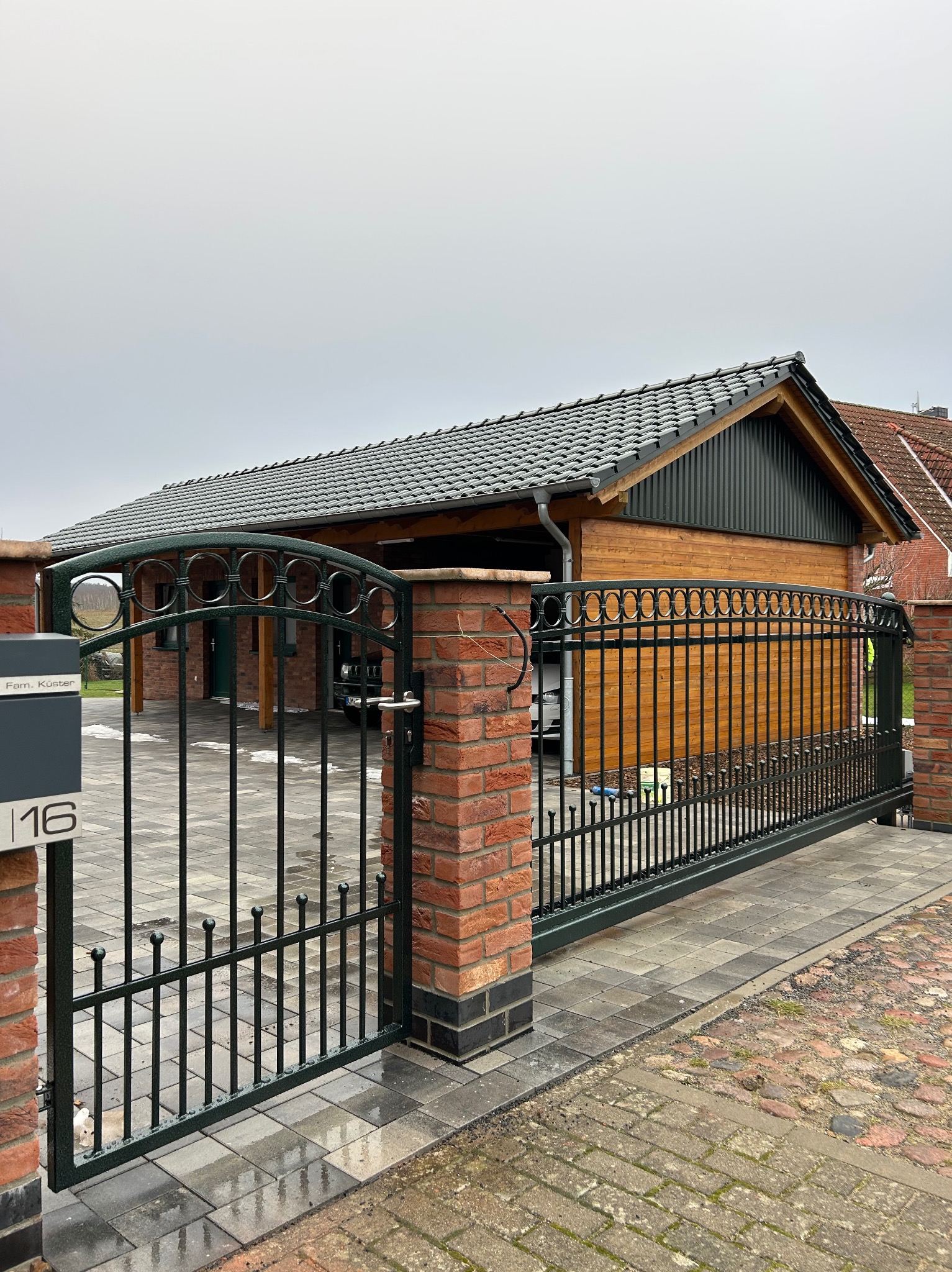 Przesuwna brama wjazdowa z metalowymi prętami i dekoracyjnymi okręgami na tle drewnianego garażu z szarą dachówką, widok z zewnątrz posesji, skrzynka pocztowa z numerem domu po lewej.